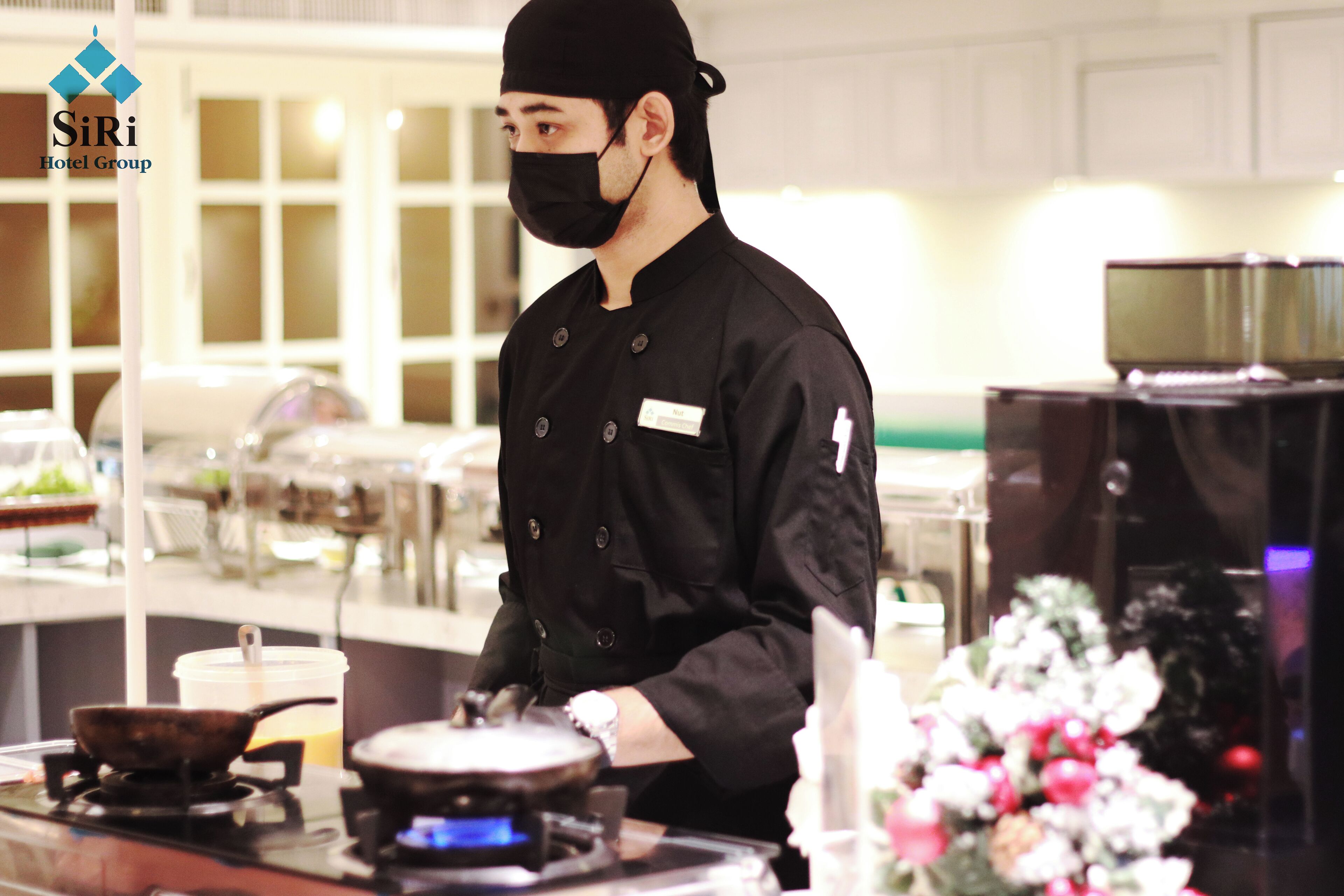 A chef in a black uniform with a name tag, cooking in a professional kitchen with stainless steel appliances and a white countertop.