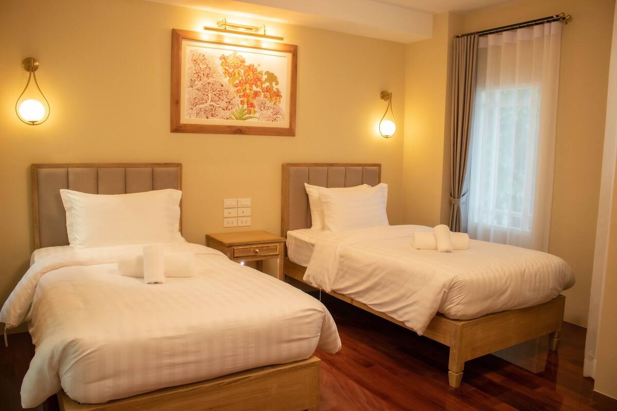 A hotel room with two beds, a nightstand, a framed floral artwork, and a window with curtains.