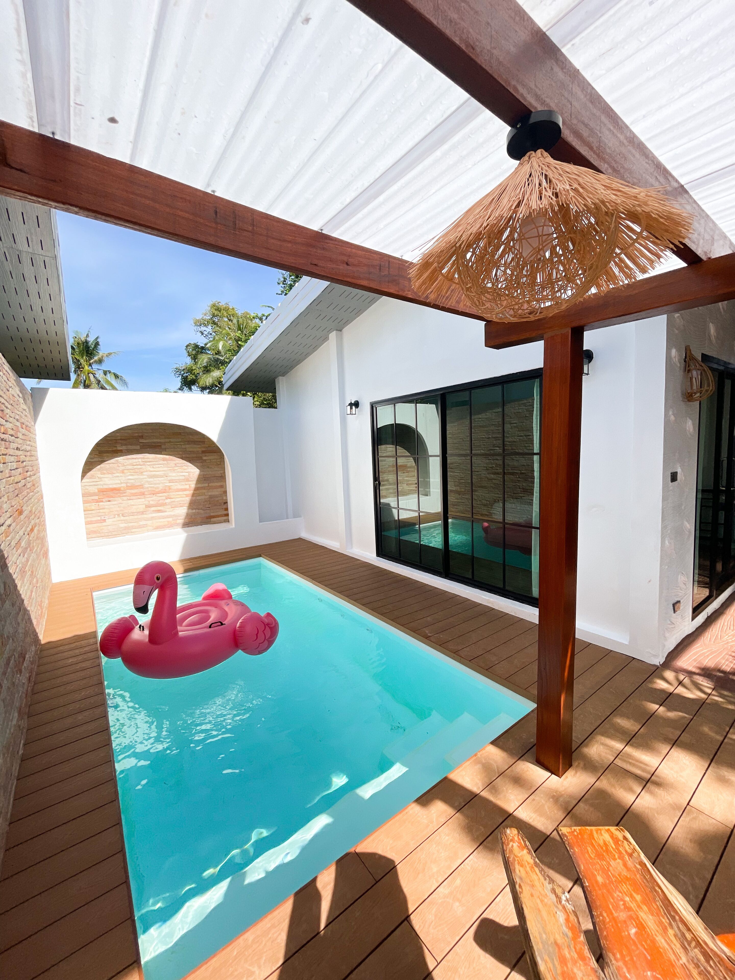 A swimming pool with a pink flamingo float, wooden deck, and a covered area with a woven ceiling.