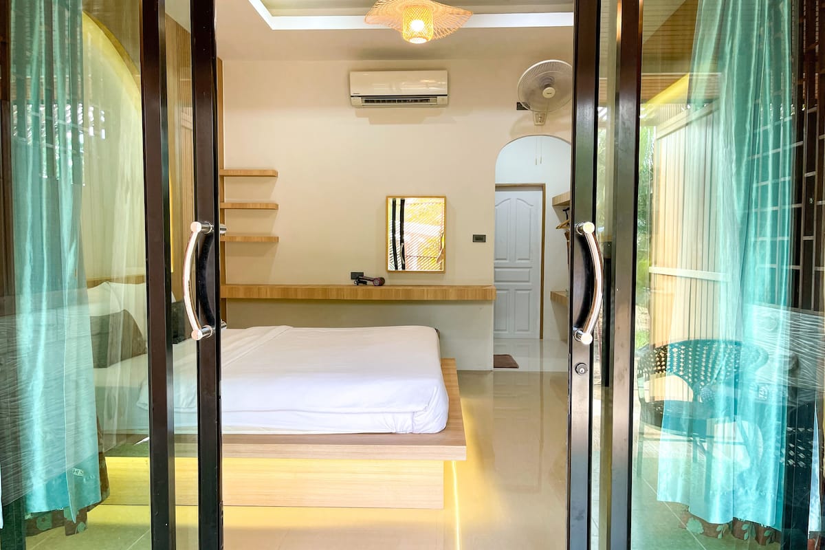 A modern hotel room with a bed, a small table, and a chair. There is a door leading to another room and a window with curtains.