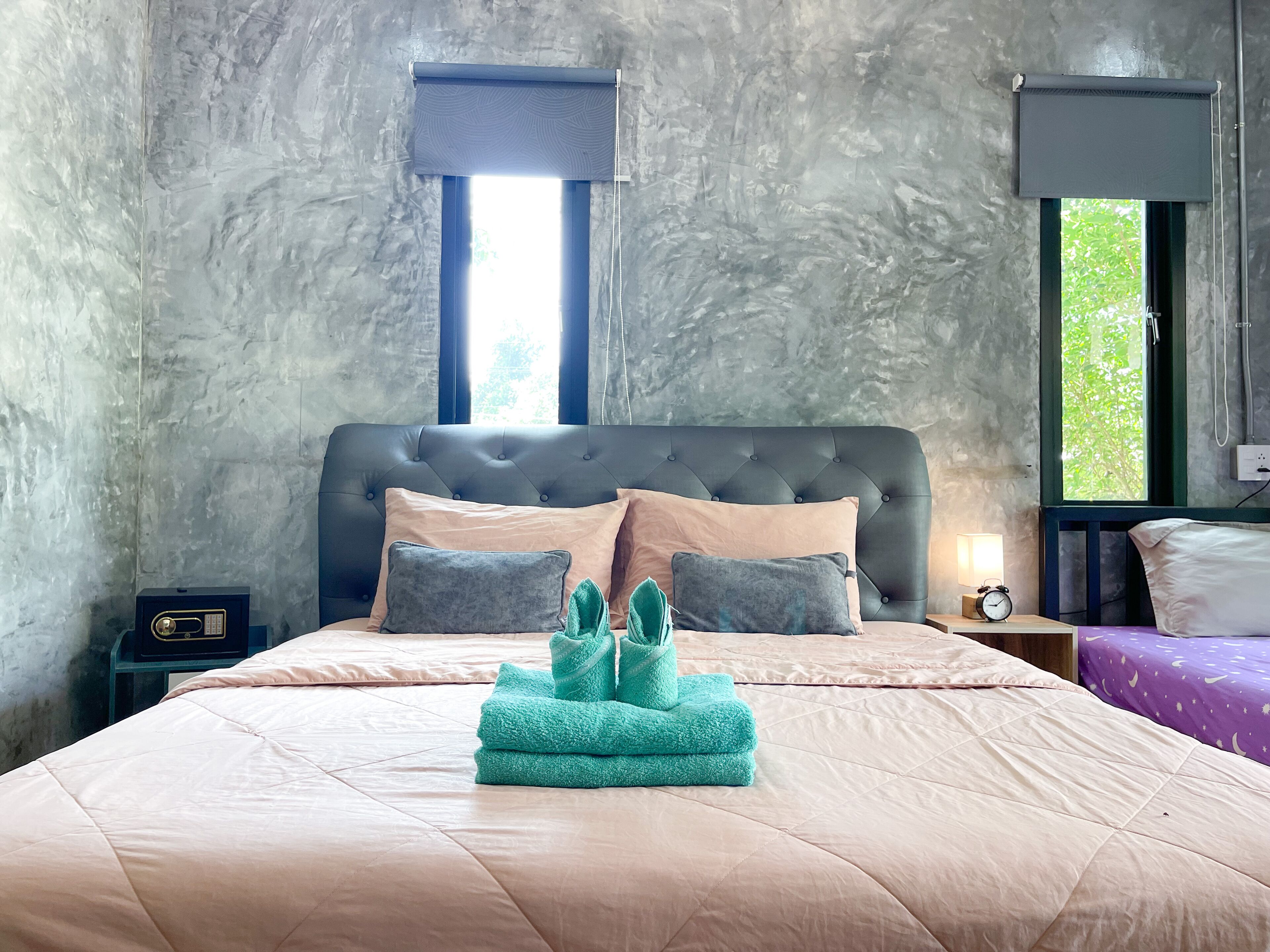 A bedroom with a tufted headboard, a bed with a beige quilt, two pillows, a nightstand with a lamp, and a window with a view of greenery.