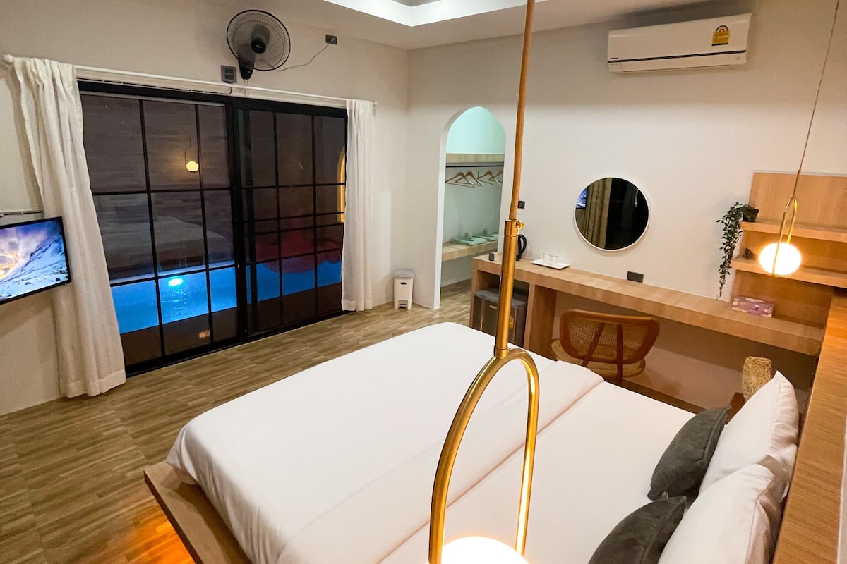 A modern hotel room with a large bed, a desk with a chair, a mirror, and a view of the sea through a sliding door.
