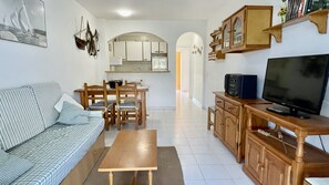 Una sala de estar con sofá, mesa de centro, televisión y un área de cocina con armarios y mesa de comedor.