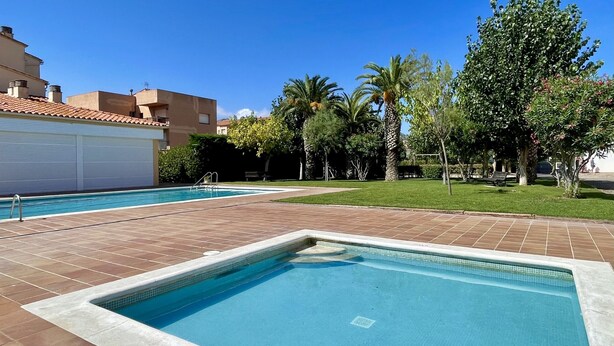 A swimming pool with a tiled deck, surrounded by greenery and a building with a closed garage door. - ESPLENDID 11 (Estartit)
