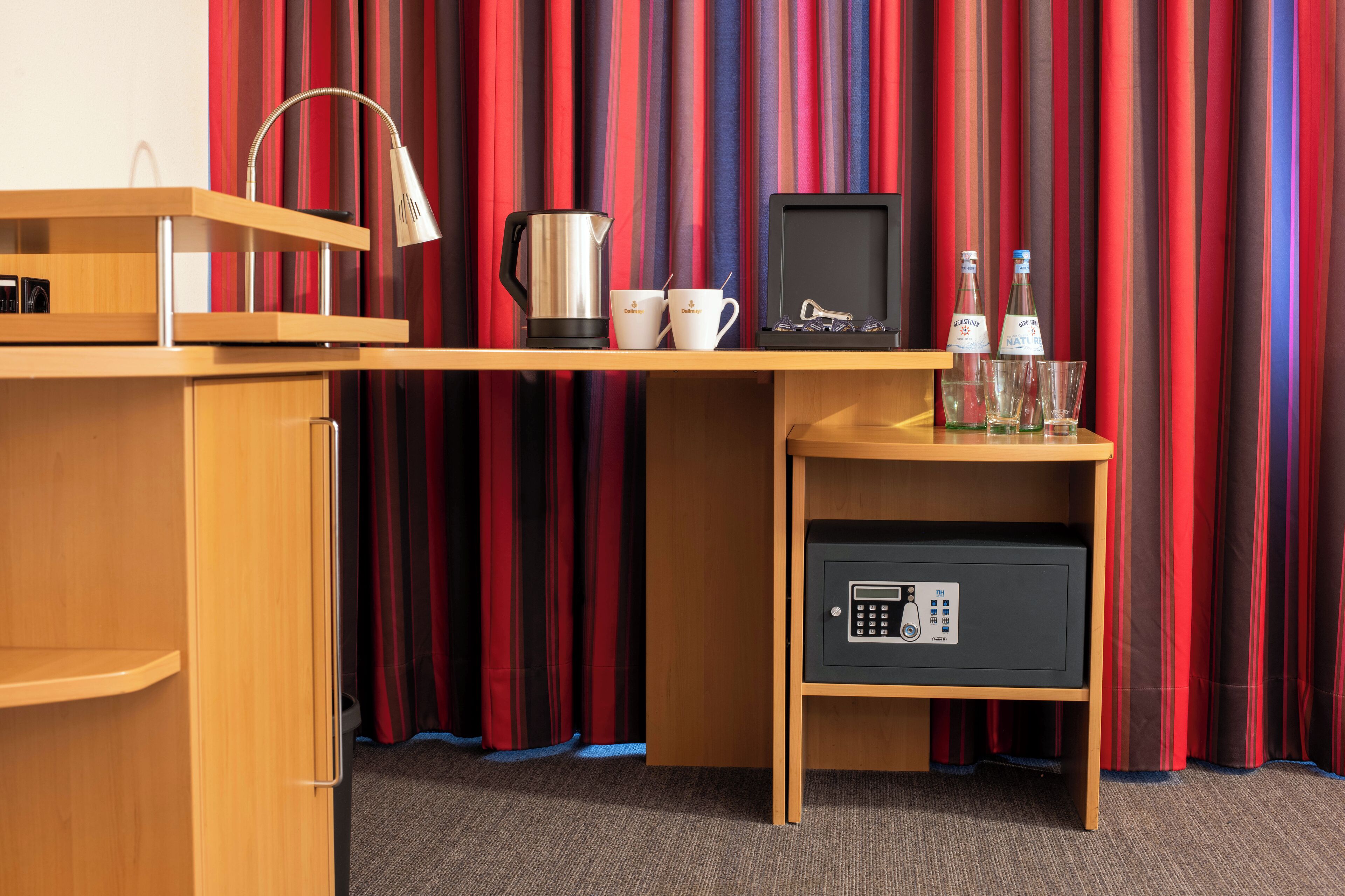 A hotel room with a wooden desk, a coffee maker, two mugs, a safe, and a pair of glasses.