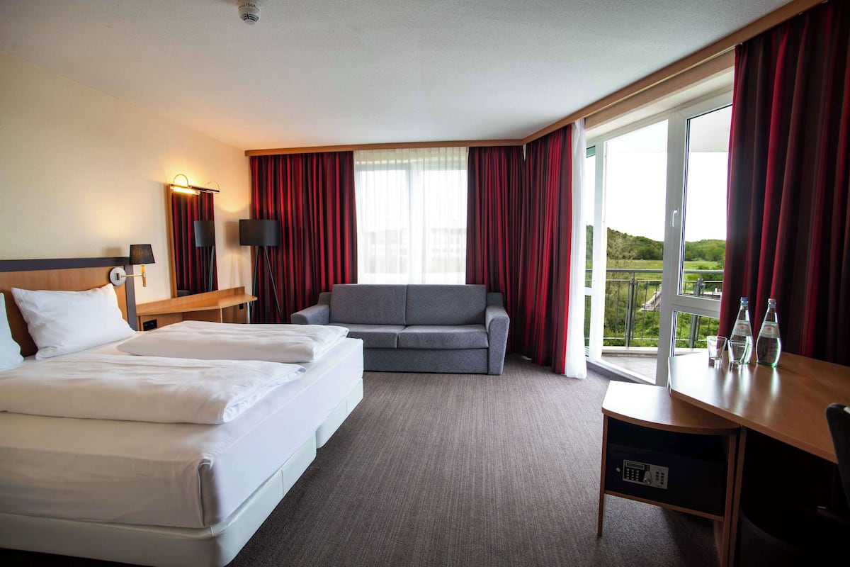 Une chambre d’hôtel avec deux lits, un canapé, un bureau avec une chaise et une grande fenêtre avec des rideaux.