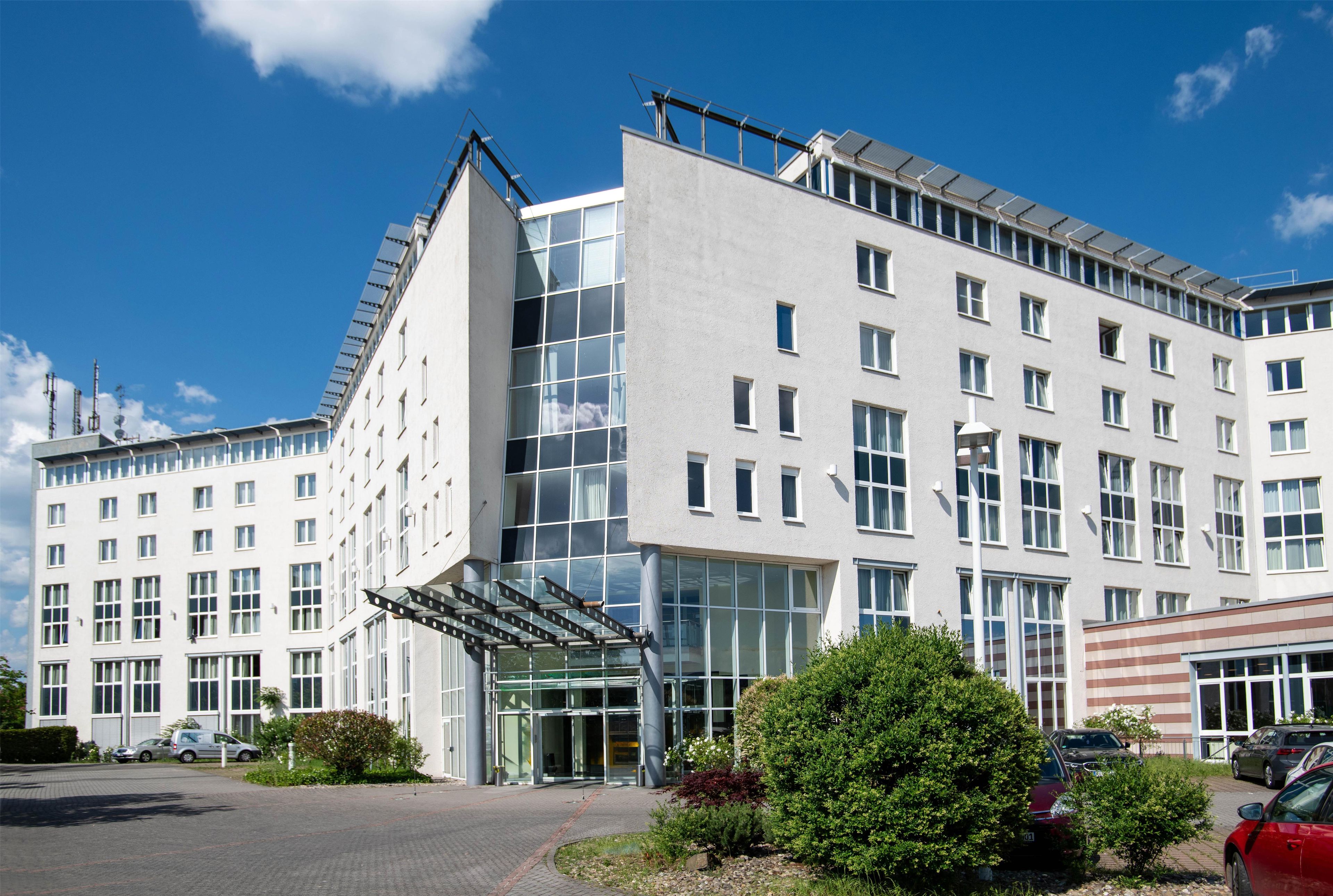 A modern building with a glass entrance, multiple windows, and a parking area with cars.