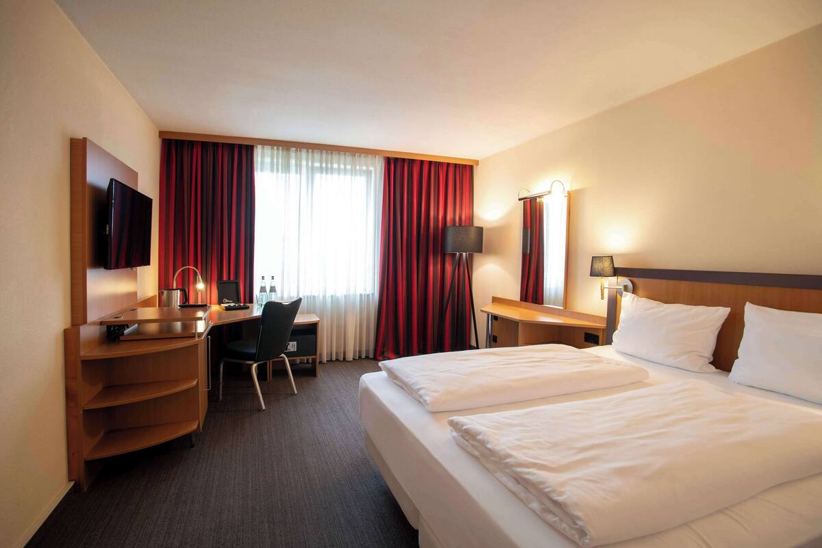 Une chambre d’hôtel avec deux lits, un bureau, une chaise, une télévision et une fenêtre avec des rideaux.