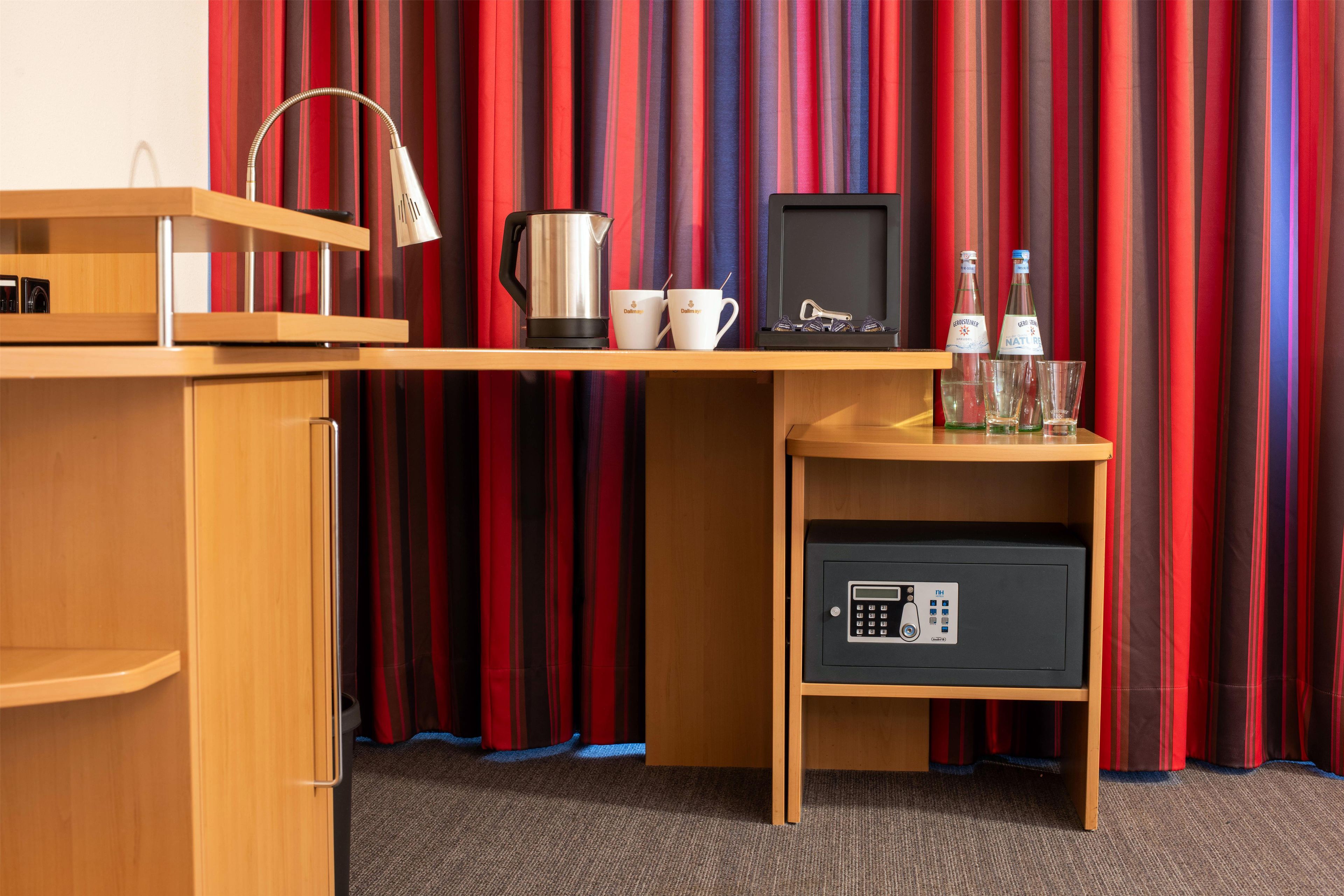 A hotel room with a wooden desk, a coffee maker, a cup, two bottles, and a safe.