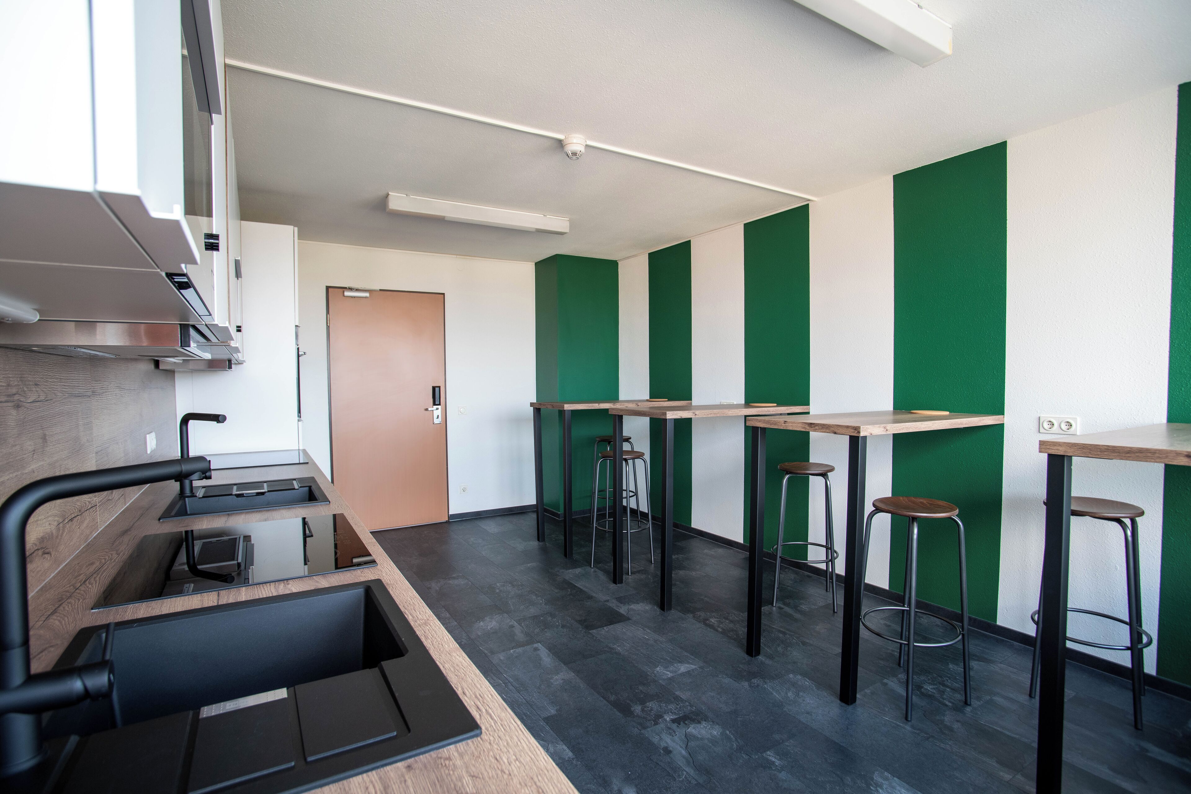 A modern kitchen with a built-in oven, a sink, and a wooden countertop. There are three stools and a table with a green accent wall.