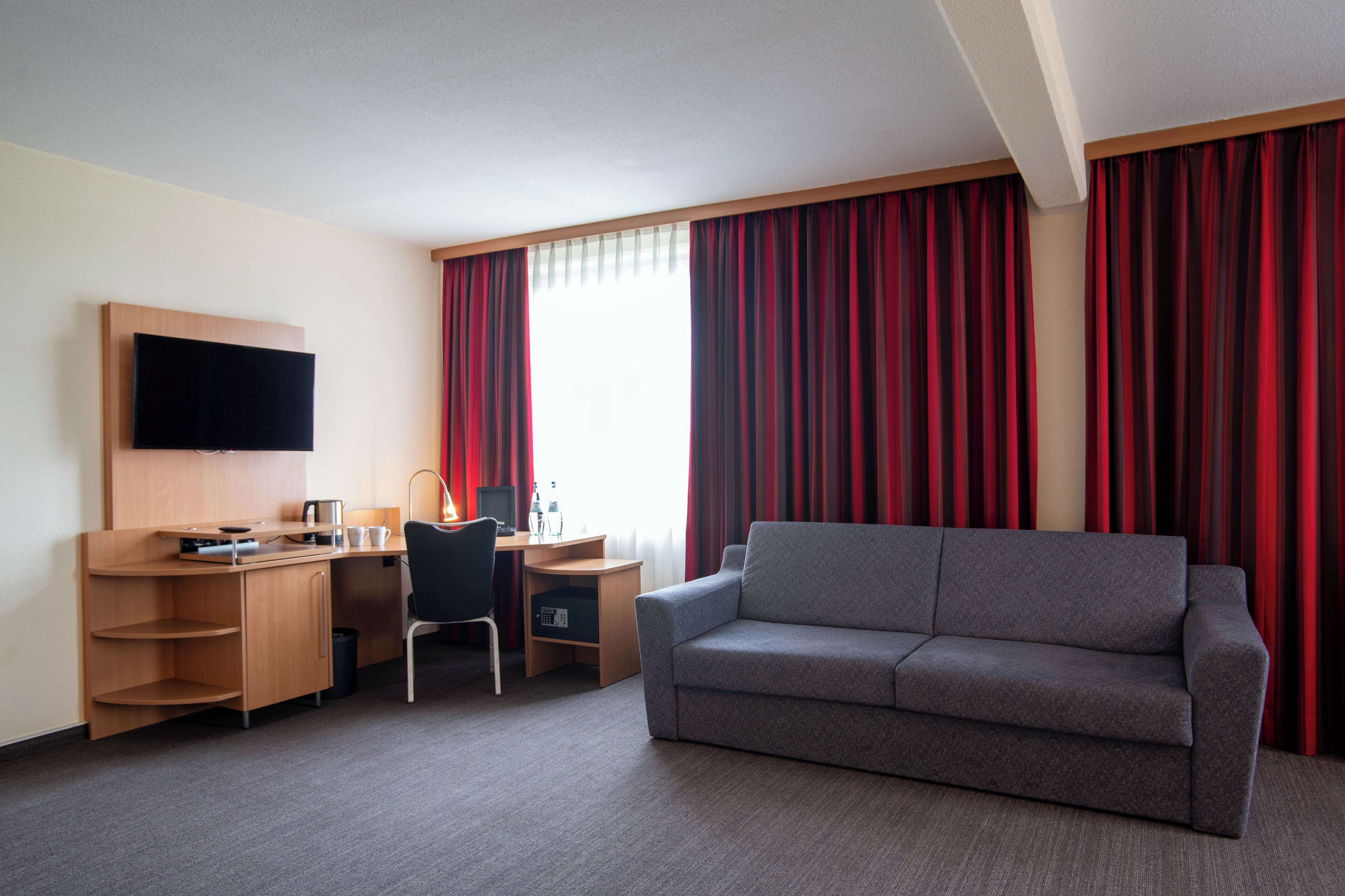 Een hotelkamer met een grijze bank, een houten bureau met een flatscreen televisie, een stoel en een raam met rode gordijnen.