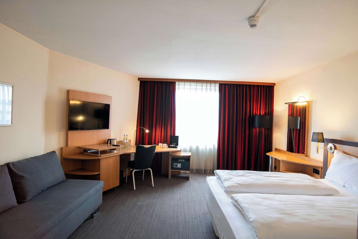 Une chambre d’hôtel comprenant un lit, un canapé, un bureau avec un ordinateur, une télévision et une fenêtre avec des rideaux.