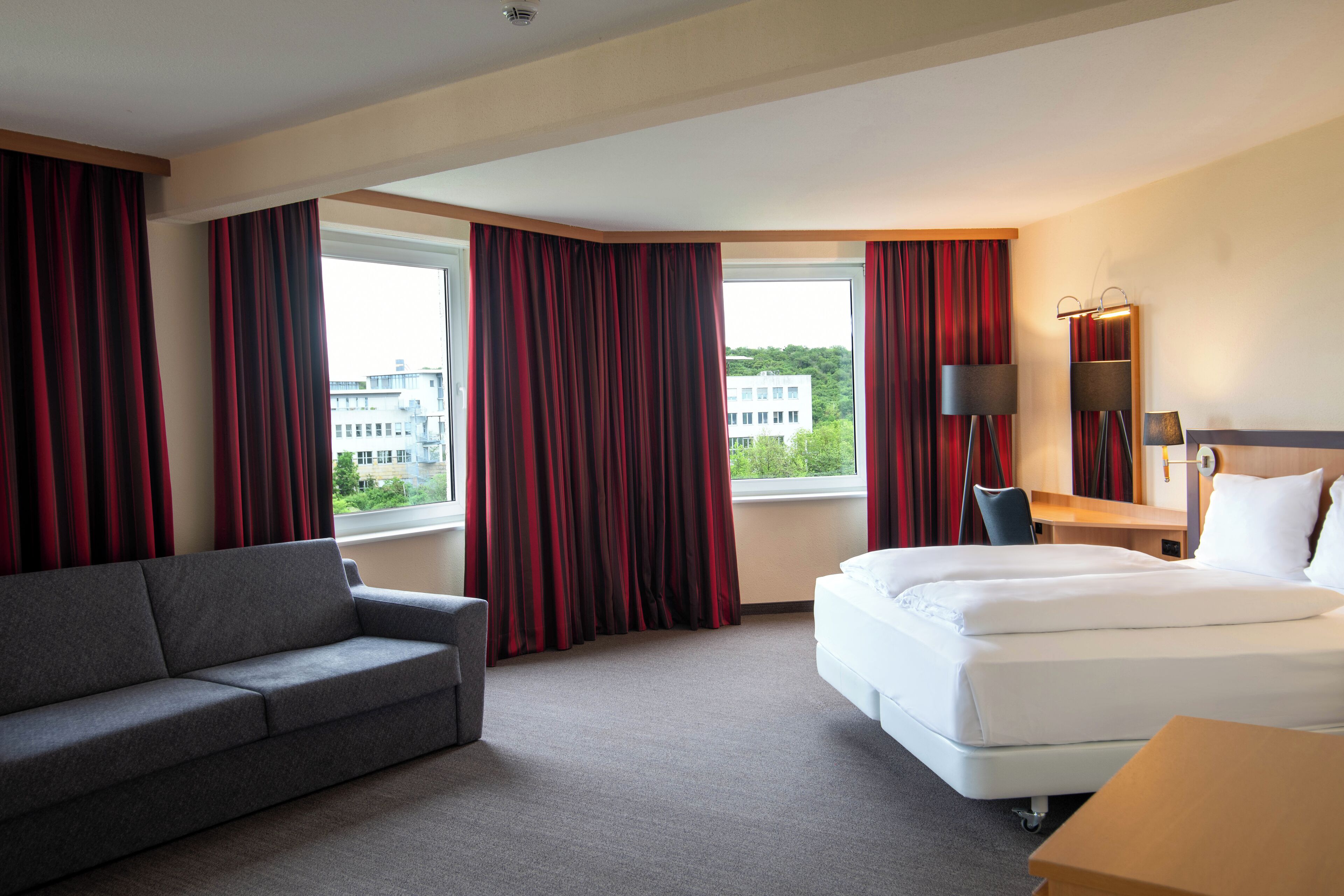Habitación de hotel con cama, sofá, escritorio y vistas a edificios a través de amplias ventanas.