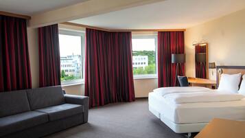 Habitación de hotel con cama, sofá, escritorio y vistas a edificios a través de amplias ventanas.
