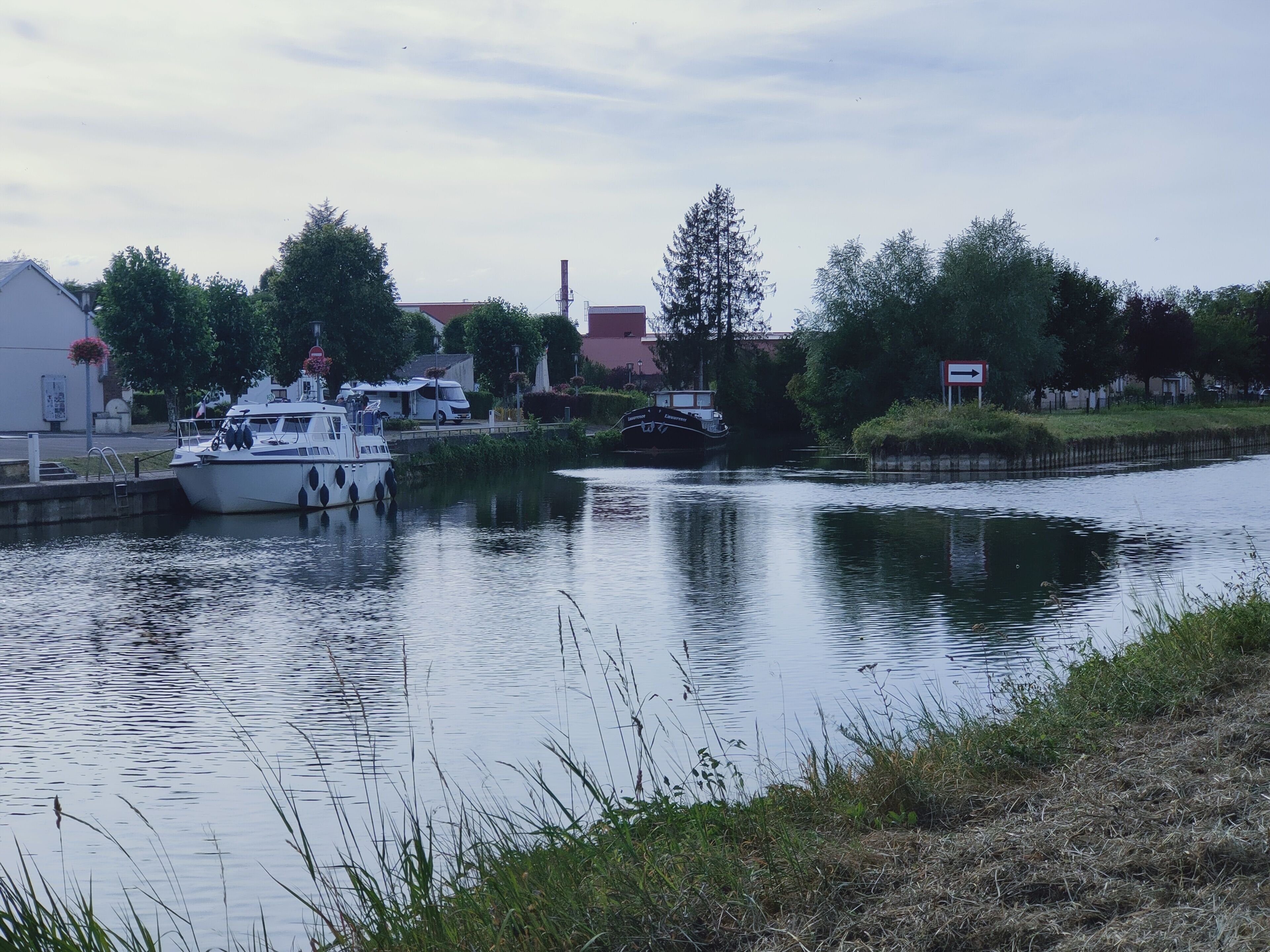 Een rustige rivier met stilgelegde boten aan de linkerkant, omgeven door begroeiing en gebouwen.