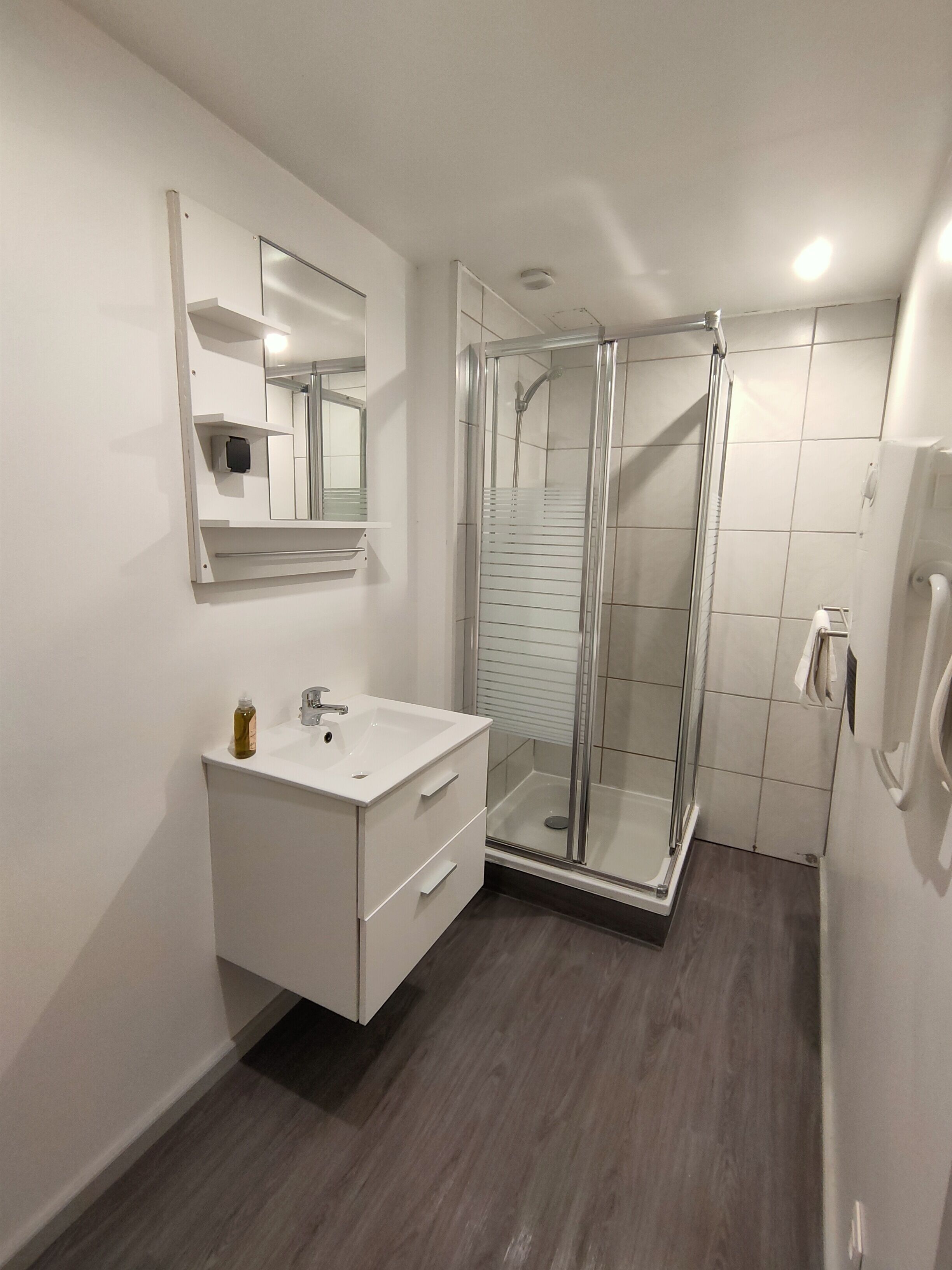 Casa de banho moderna com cabine de chuveiro em vidro, toucador branco com gavetas, espelho e dispensador de sabonete fixado na parede.