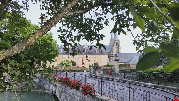 A stone bridge over a river with red flowers, surrounded by greenery and a historic building with a tower in the background.