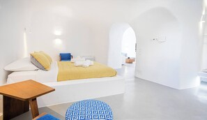 Una habitación de hotel moderna con una cama, una pequeña mesa, un reposapiés azul estampado y vistas a través de un arco hacia otra habitación.