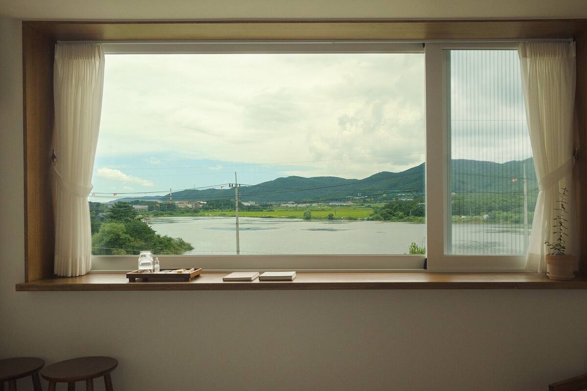 A room with a large window overlooking a river and hills. There is a wooden shelf with a tray, a notebook, and a potted plant.