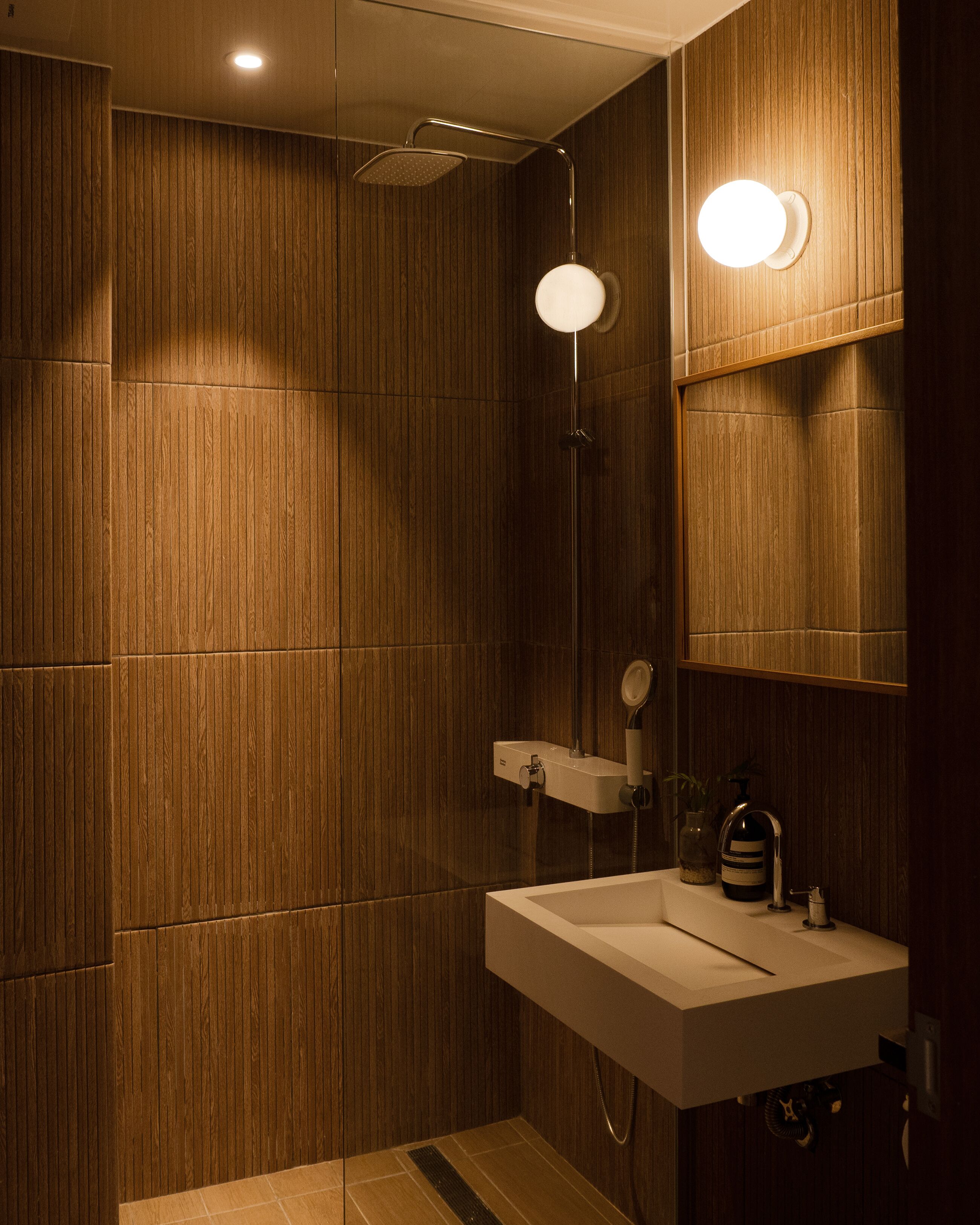 A modern bathroom with wooden paneling, a shower with a handheld showerhead, a sink with a mirror, and a wall-mounted light fixture.