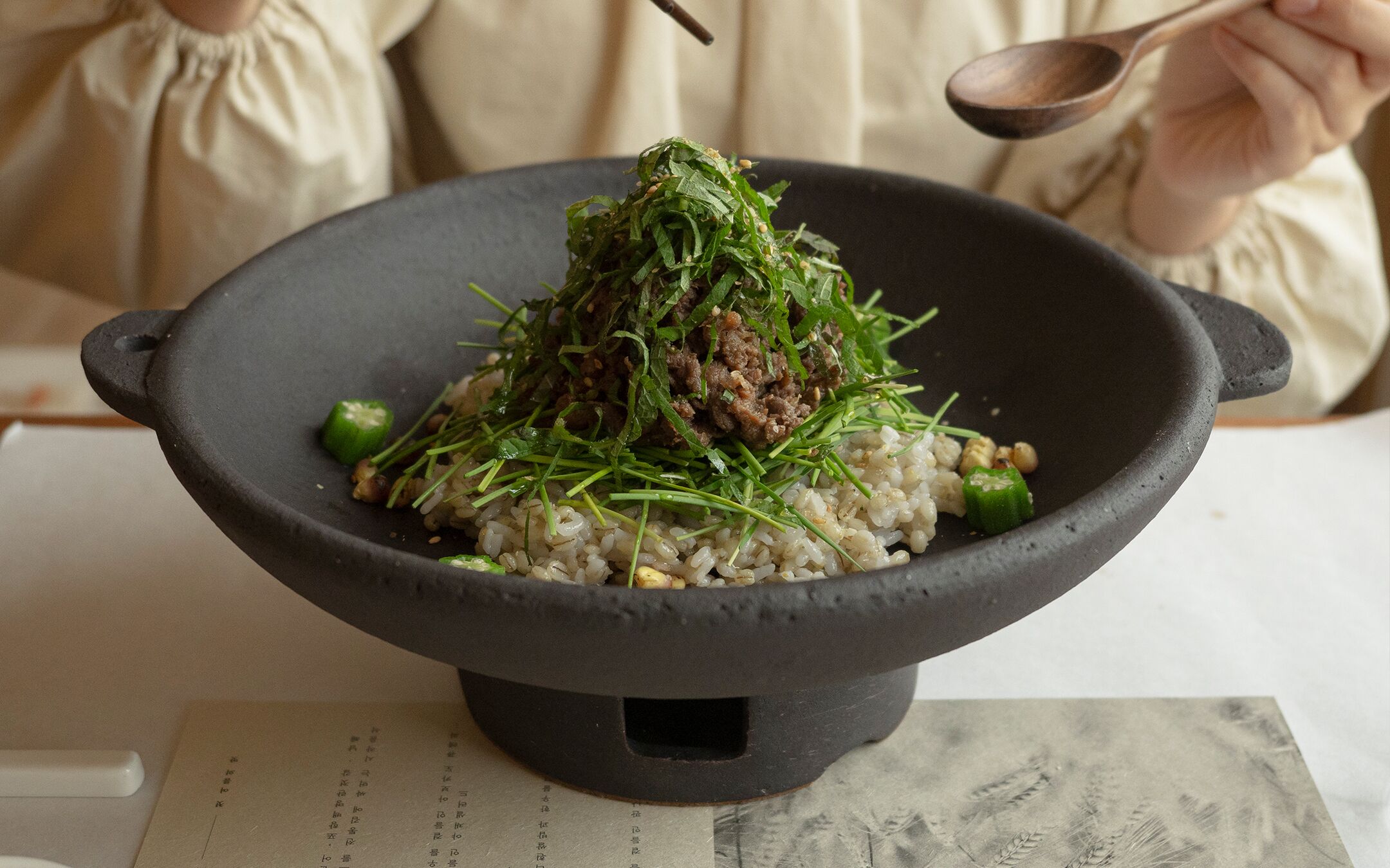 A bowl of rice topped with greens and meat, held by a person.
