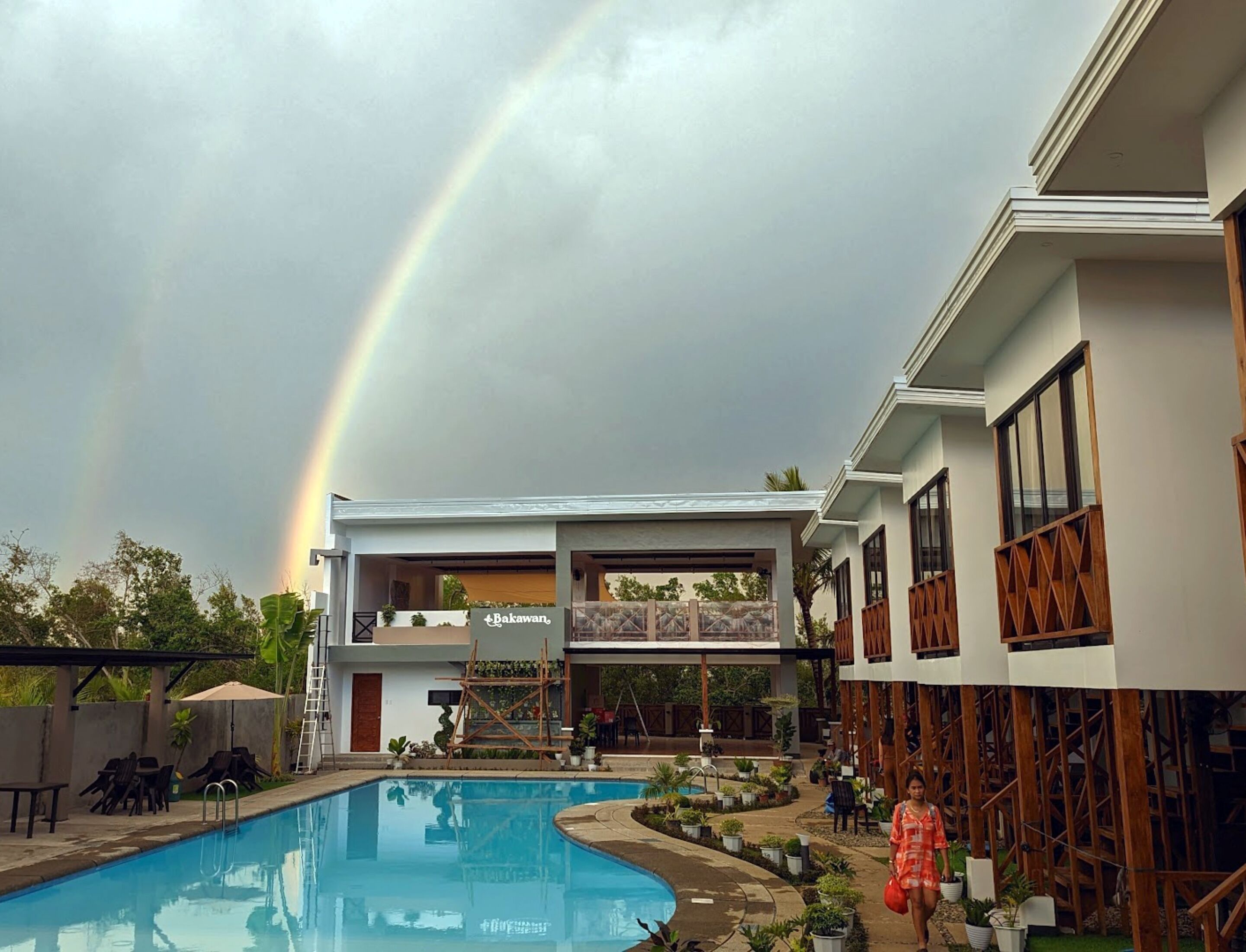 A swimming pool with a rainbow, a building with a sign reading "Bakawan", and outdoor seating.