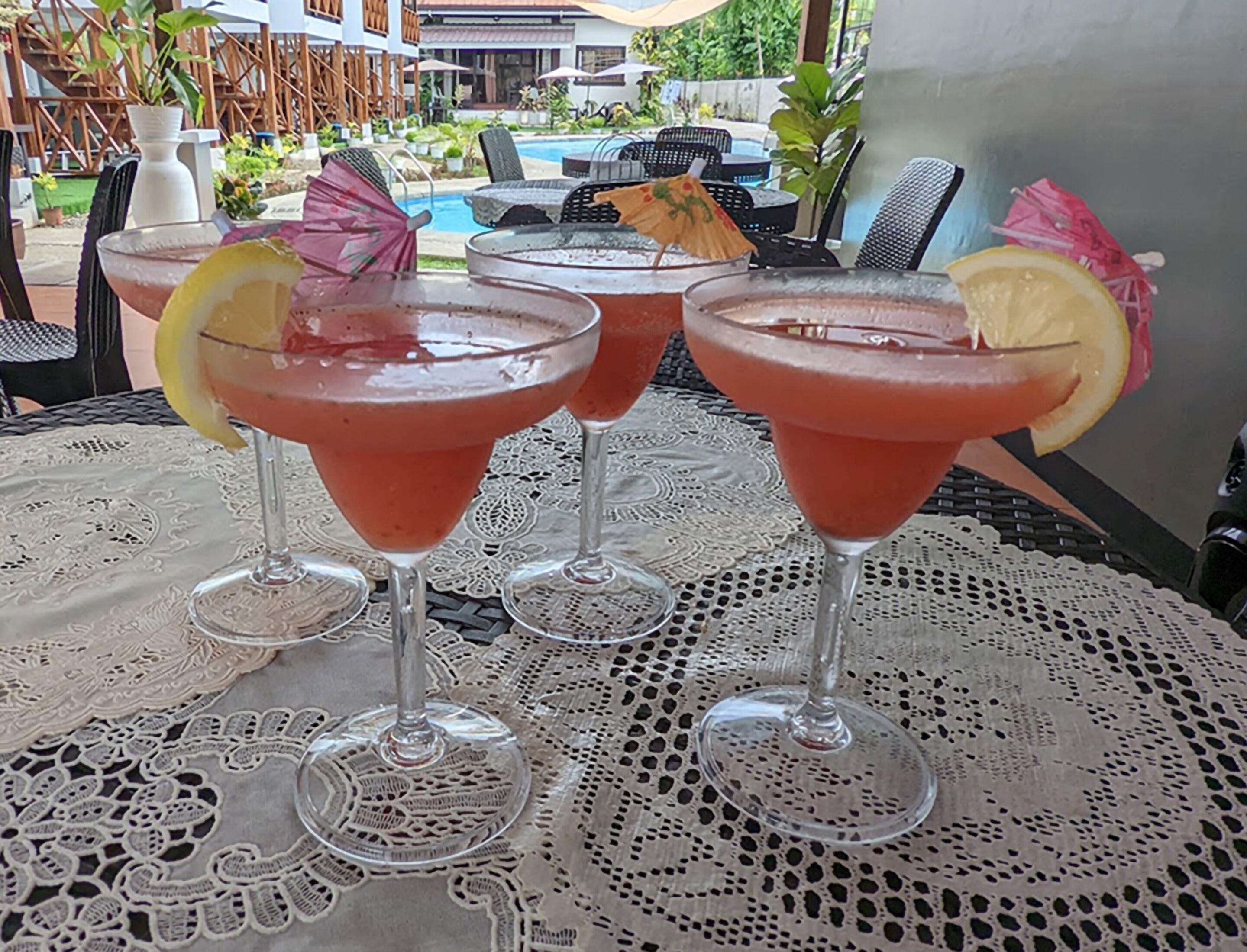 Drei Cocktails auf einem Tisch mit Spitze, umgeben von Außenplätzen und einem Pool.