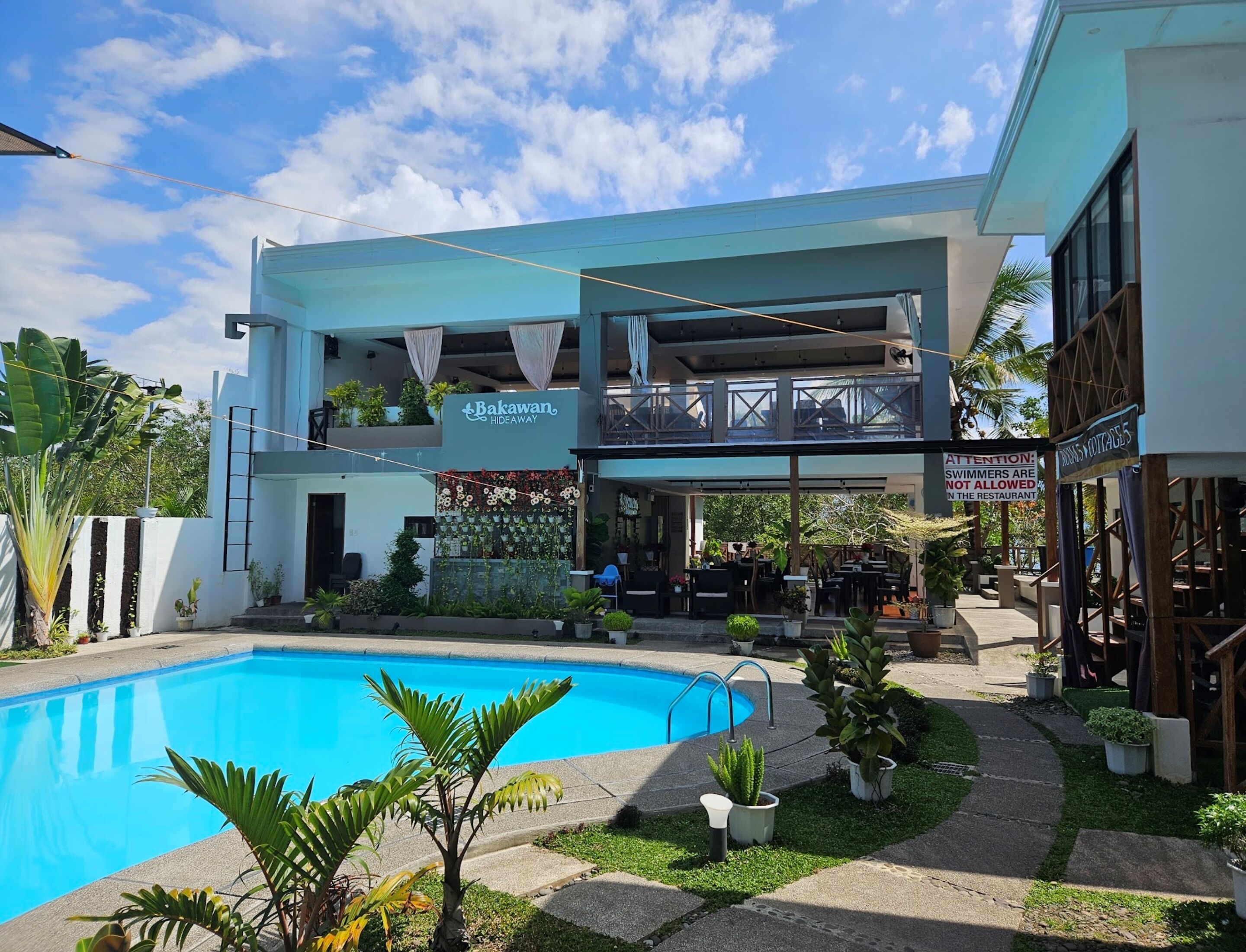Un edificio de dos pisos con un letrero que dice 'Bakawan Hideaway'. Hay un área de piscina con una piscina azul cristalina y follaje circundante.