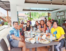 Un grupo de personas cenando en un restaurante al aire libre con terraza cubierta, mobiliario de mimbre y una variedad de platos sobre la mesa.