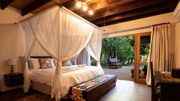 A bedroom with a canopy bed, a bench with a colorful design, a small table, and a view of the outdoors.