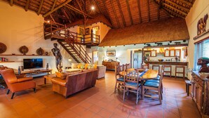 A spacious living area with a high ceiling, a staircase, a dining area with wooden furniture, and a kitchen visible in the background.