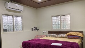 Quarto com cama de madeira, roupa de cama roxa, duas janelas com persianas padronizadas e um aparelho de ar condicionado.