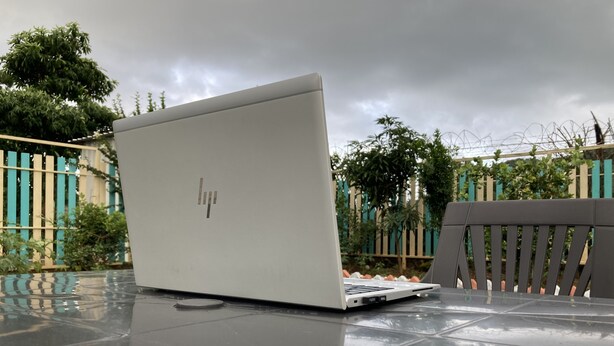 Um laptop sobre uma superfície refletora ao ar livre, com uma cerca e árvores ao fundo.