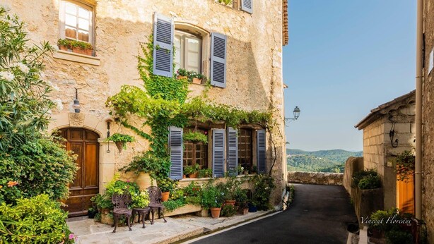 Una calle estrecha con un edificio de piedra que tiene una puerta de madera, contraventanas y macetas con plantas.