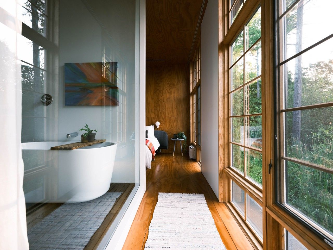 A modern bathroom with a freestanding bathtub, a wooden floor, and a large window offering a view of the outdoors.
