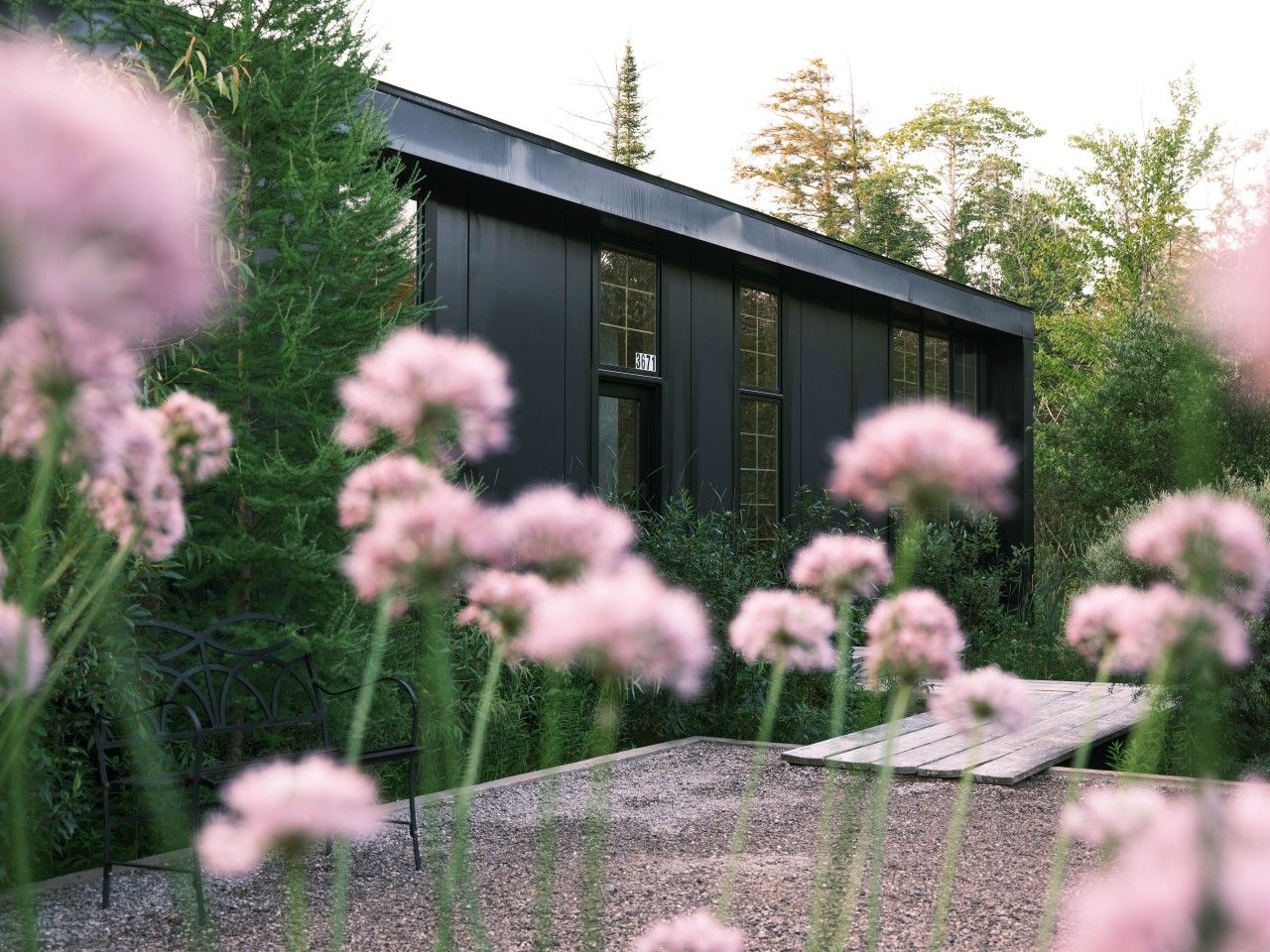 Un edificio moderno con fachada oscura, rodeado de abundante follaje y flores rosadas.