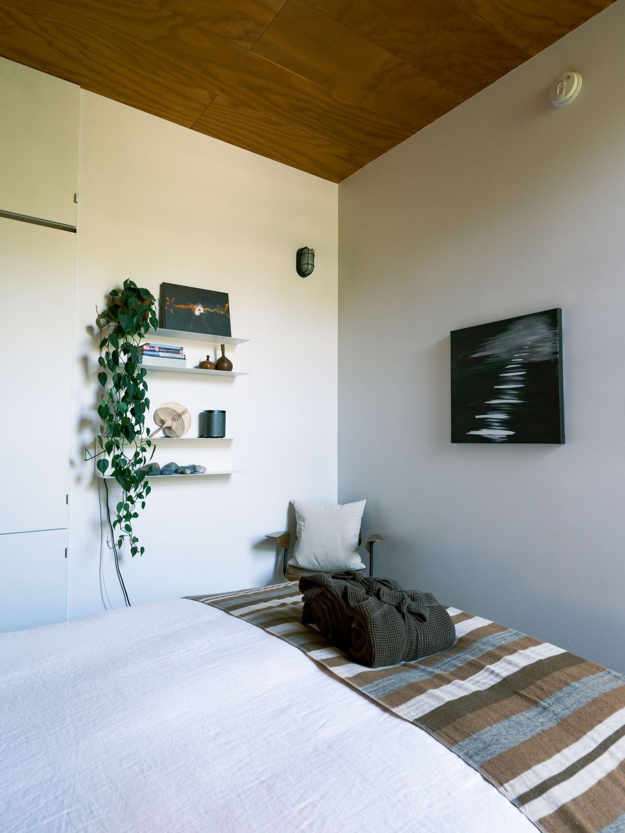 A modern bedroom with a bed, a chair, a wall-mounted shelf with books and decorative items, a potted plant, and a painting.