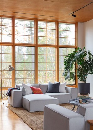 Ein modernes Wohnzimmer mit Ecksofa, großen Fenstern, einer Stehlampe und einem Blumentopf.
