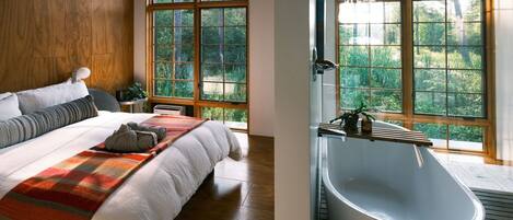 A bedroom with a large bed, a window with a view of greenery, and a modern bathtub.