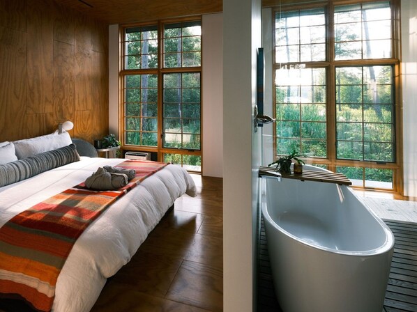 A bedroom with a large bed, a window with a view of greenery, and a modern bathtub.
