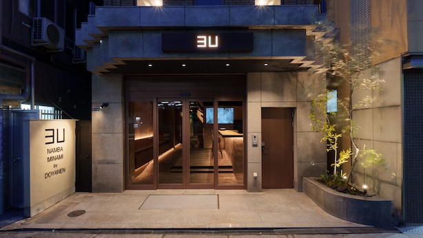 A modern building entrance with a glass door, a sign above the door, and a small garden area with plants. - 3U NAMBA MINAMI - DOYANEN HOTELS (Osaka)