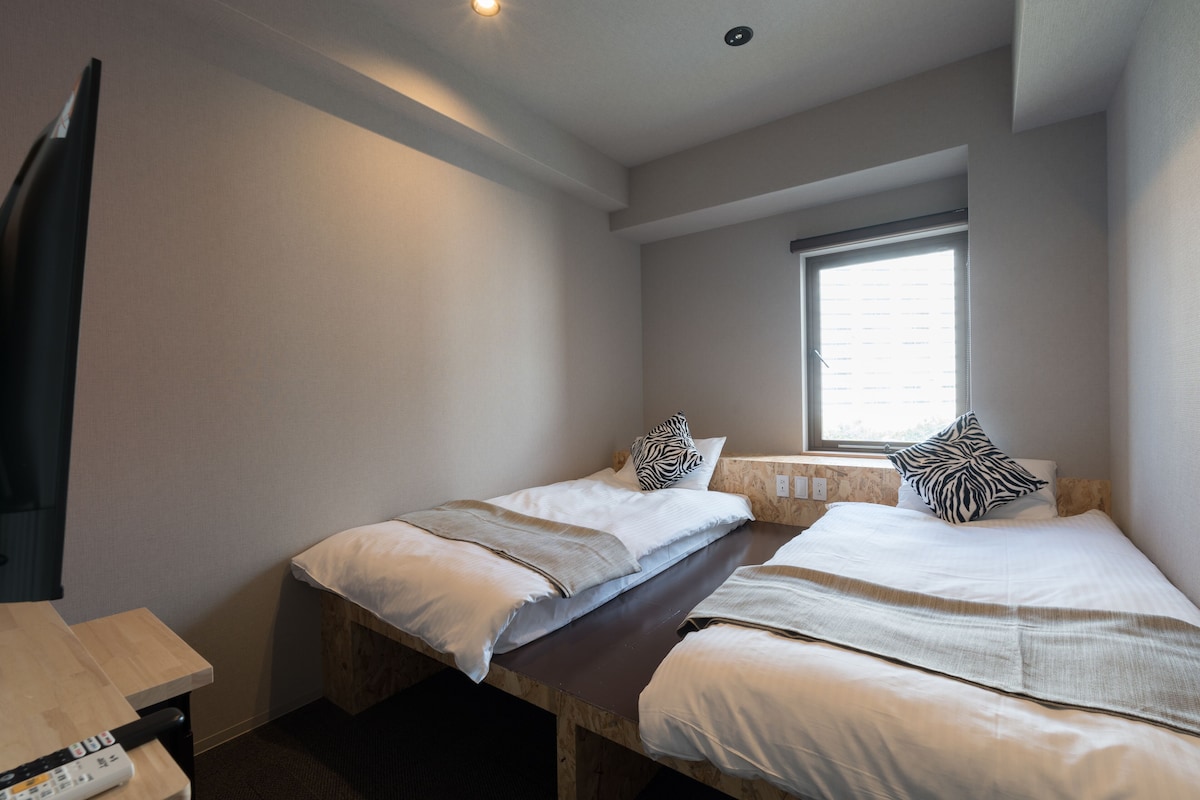 A small, modern room with two single beds, a wooden table, and a window providing natural light.