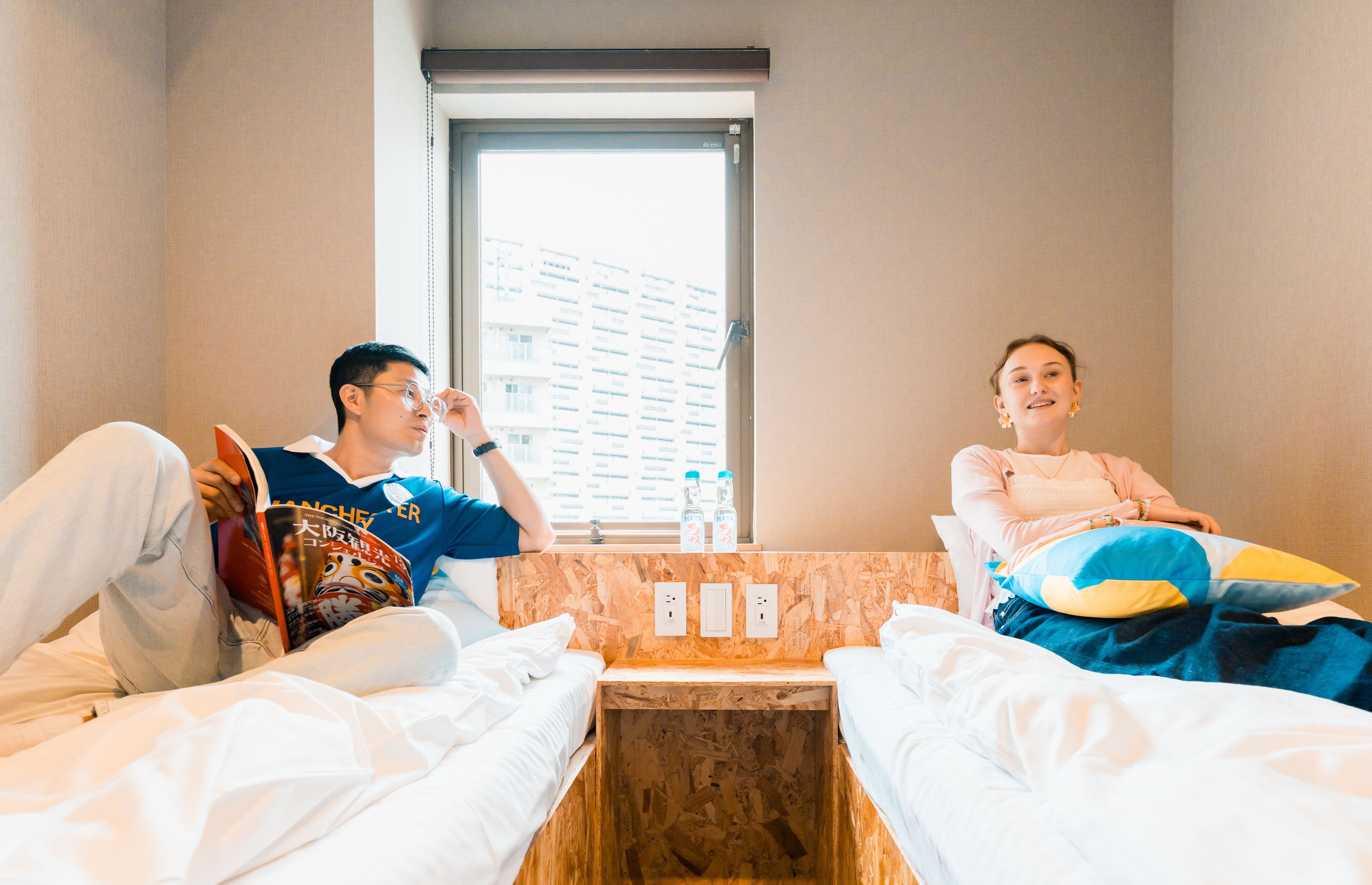 A hotel room with two beds, a wooden headboard, a window with a view of buildings, and a small table with a bottle and glasses.
