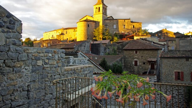 Un edificio histórico de piedra con un campanario, rodeado de arquitectura tradicional y un muro de piedra.