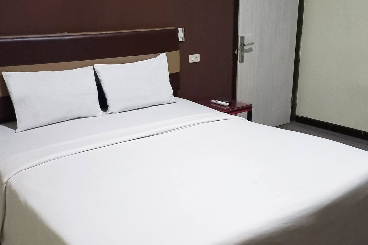 Ein ordentlich bezogenes Bett mit weißen Leinen und Kissen, ein hölzerner Kopfteil, ein kleiner Nachttisch mit Fernbedienung und eine Tür mit Griff.