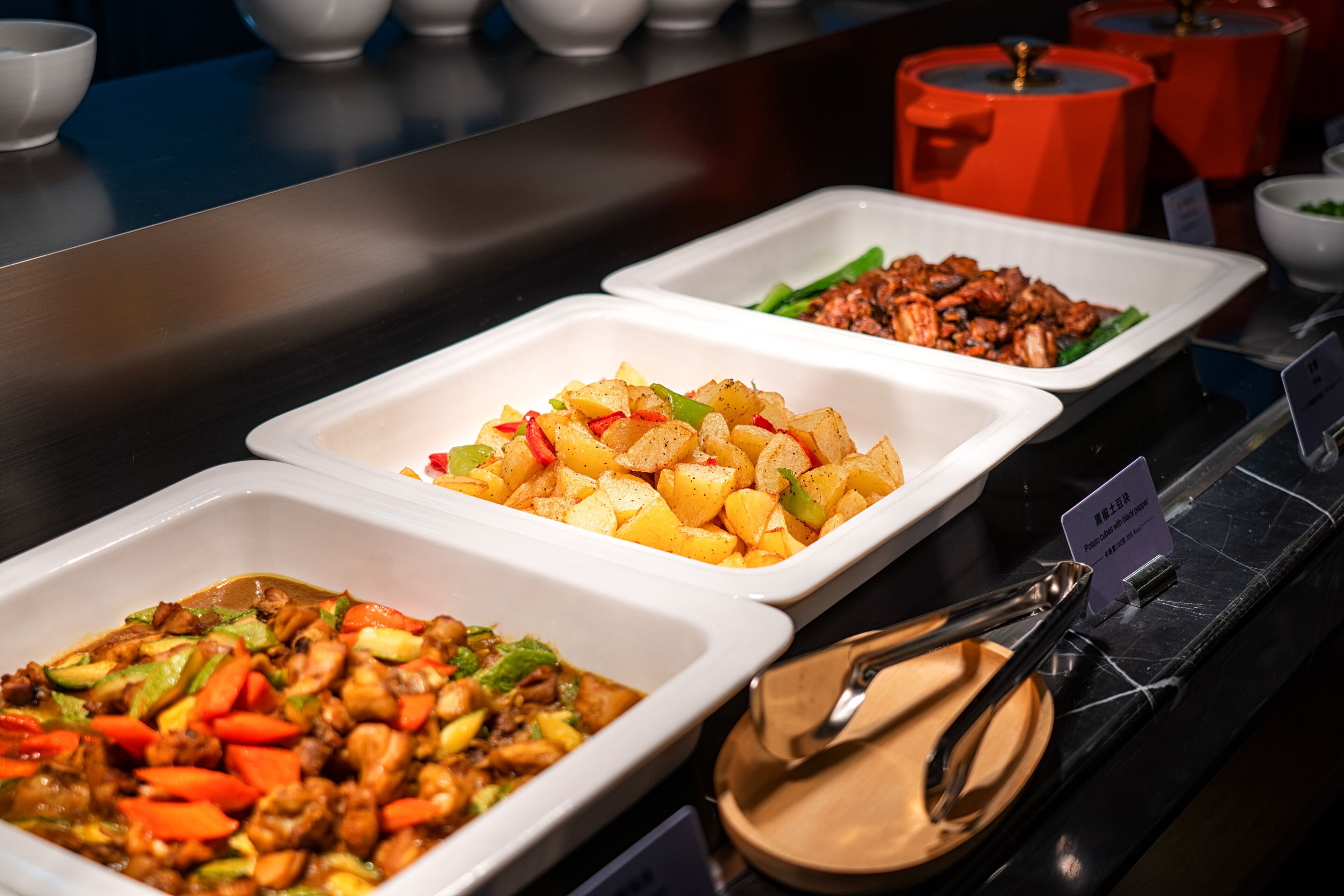A buffet with various dishes in white trays, including a vegetable stir-fry, a meat dish, and a potato dish.