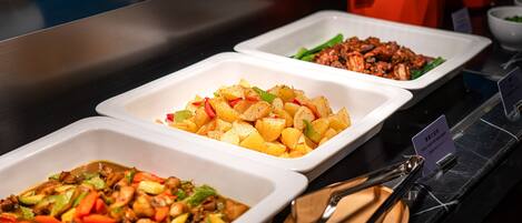 A buffet with various dishes in white trays, including a vegetable stir-fry, a meat dish, and a potato dish.