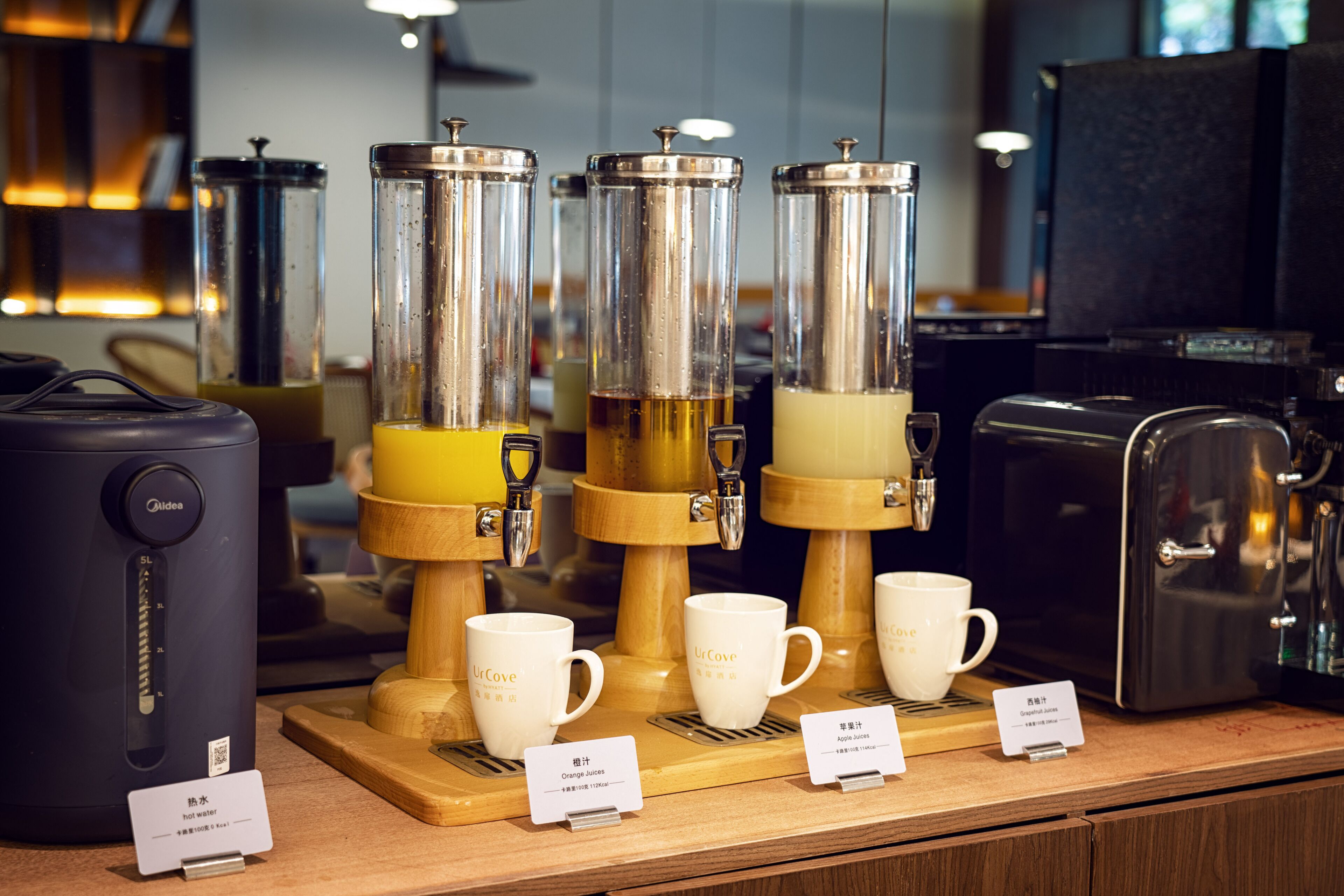 Una estación de café con un dispensador de agua caliente, dos molinillos y tazas etiquetadas para zumo de naranja y zumo de manzana.