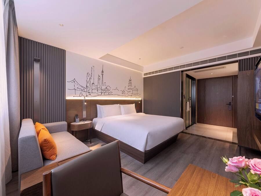 A modern hotel room with a bed, a sofa, a small table, and a wall-mounted artwork depicting a cityscape.