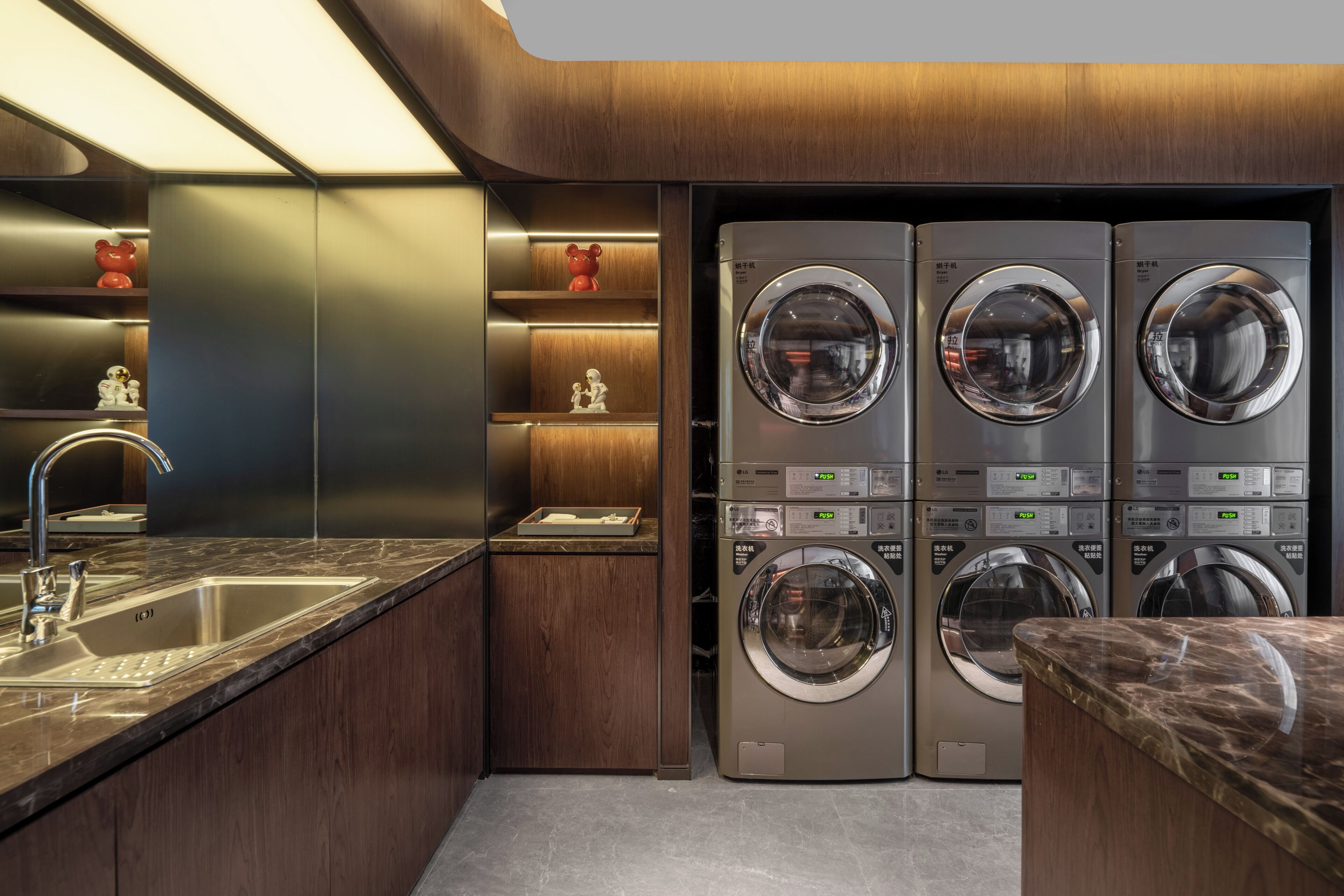 A modern laundry room with four stacked washing machines, a stainless steel sink, and wooden shelves with decorative items.
